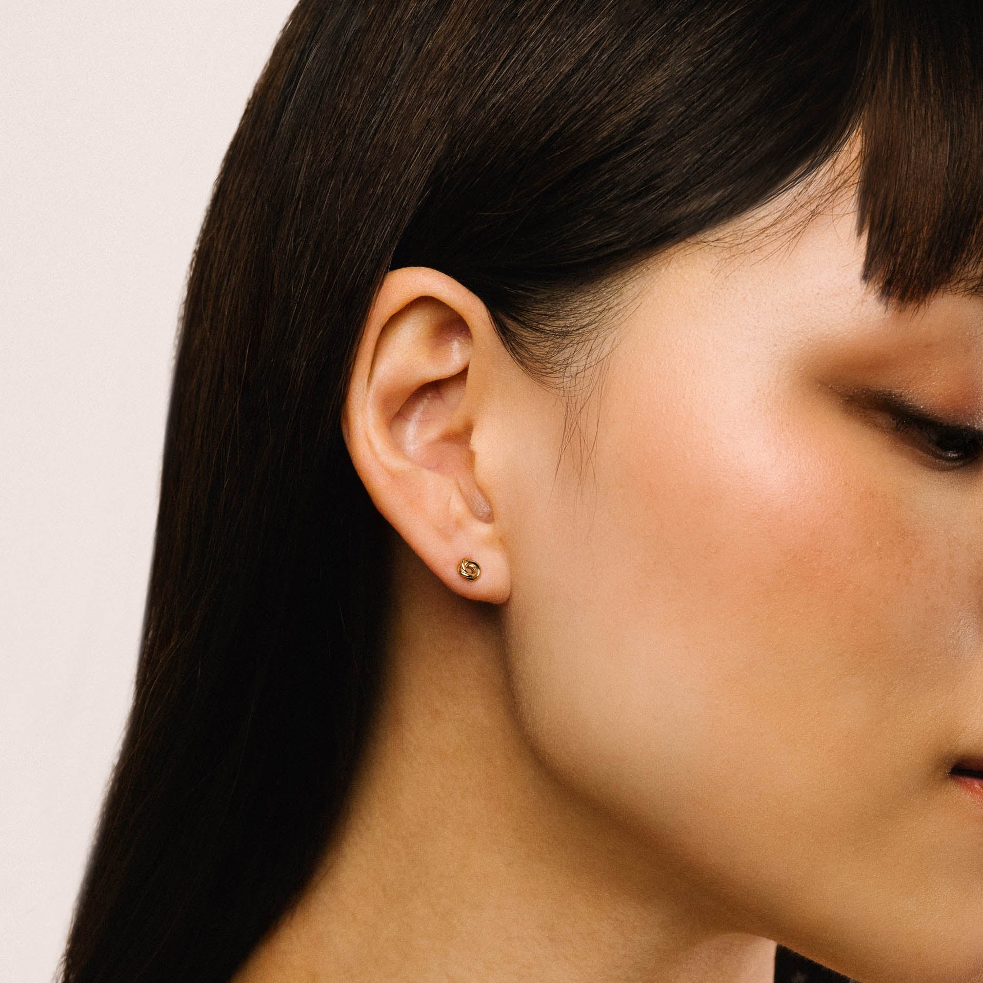 A close-up side profile of a woman wearing a small gold stud earring on her earlobe against a plain light background.
