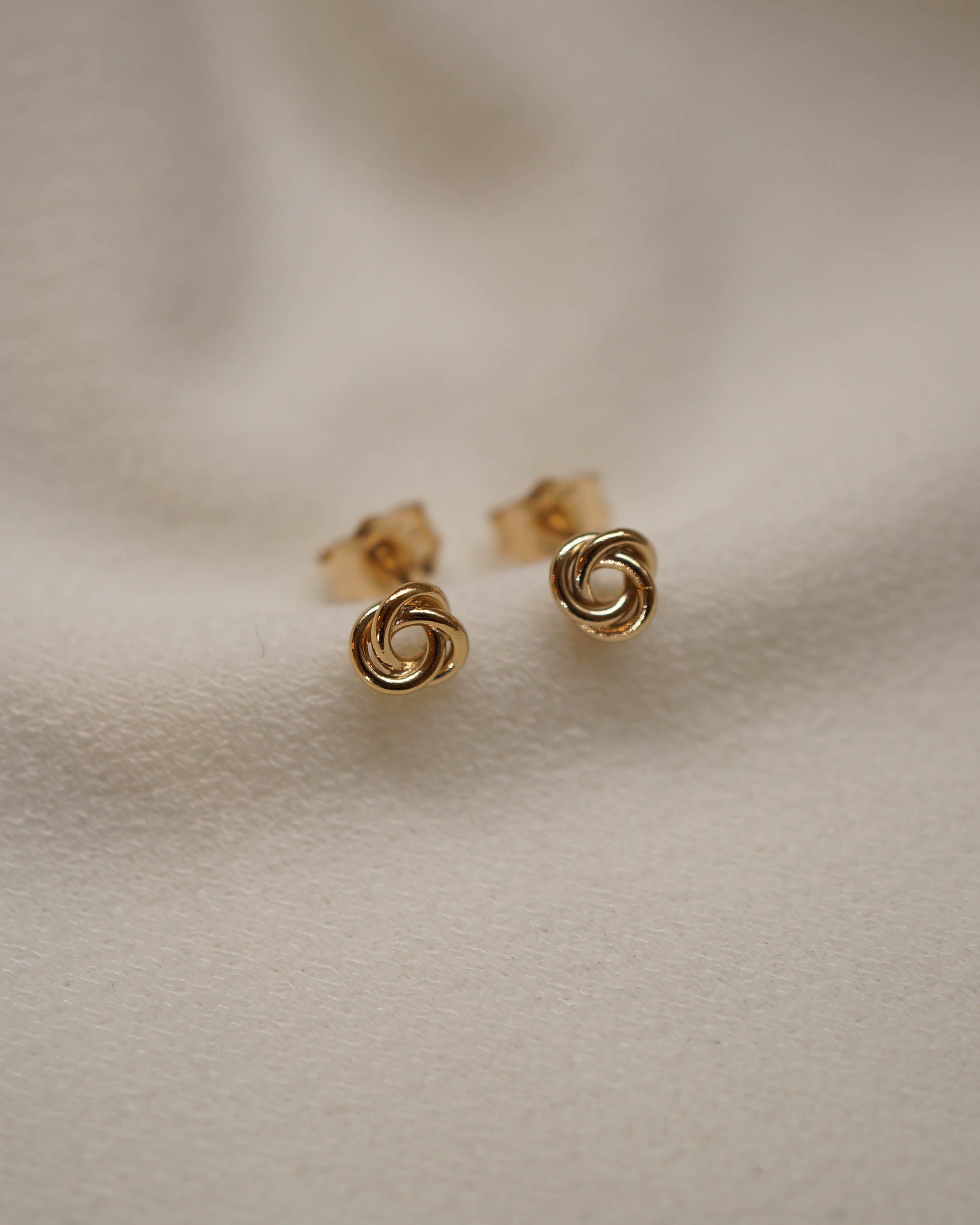 A pair of small gold earrings with a twisted knot or spiral design, resting on a soft, textured beige fabric background.